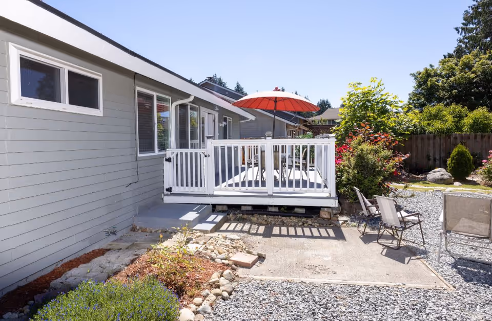 Outdoor patio area of a residential facility with a small white deck featuring a red umbrella and outdoor table and chairs. The patio is surrounded by gravel, plants, and a wooden fence with trees and bushes in the background under a clear blue sky.