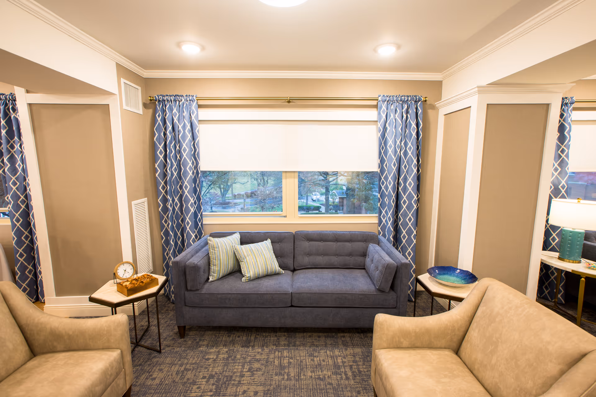 A cozy living room area with a blue sofa adorned with two striped pillows, flanked by two beige armchairs. The room features blue patterned curtains on either side of a large window with a white roller shade. There are two small side tables, one with a clock and decorative items, and the other with a blue decorative bowl. A lamp with a turquoise base is visible on the right side.