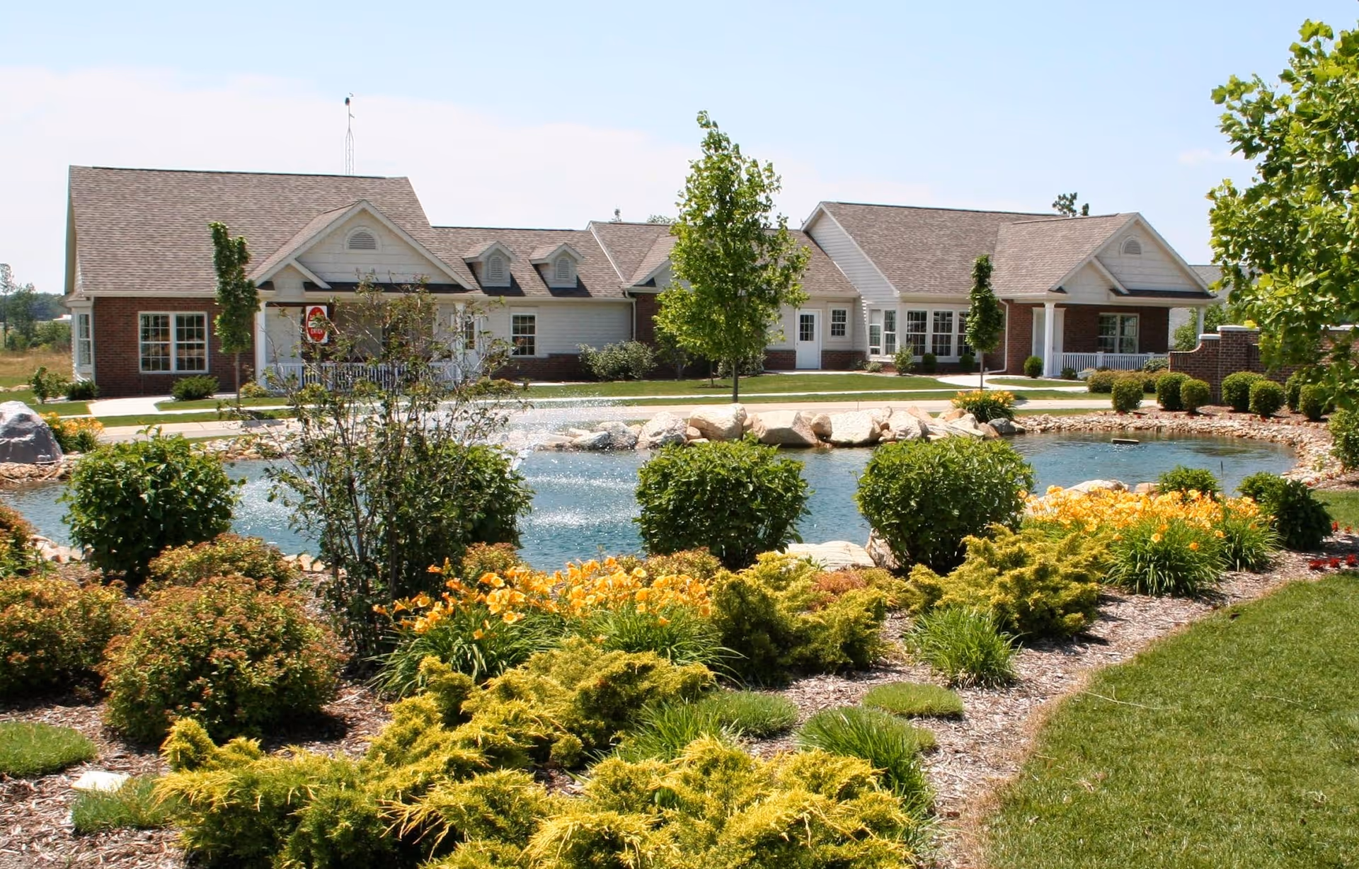 A landscaped outdoor area at Greencroft Middlebury featuring a pond with a water fountain, surrounded by bushes, flowering plants, and trees. In the background, there is a single-story building with a combination of brick and siding exterior, multiple windows, and a covered porch.