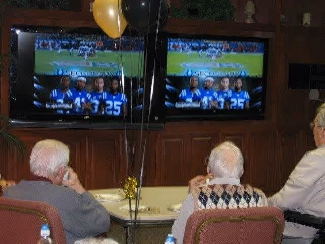 Three elderly men sitting at a table watching two large televisions mounted on a wooden wall, showing a football game and players in blue jerseys. There are black and gold balloons tied to the table.
