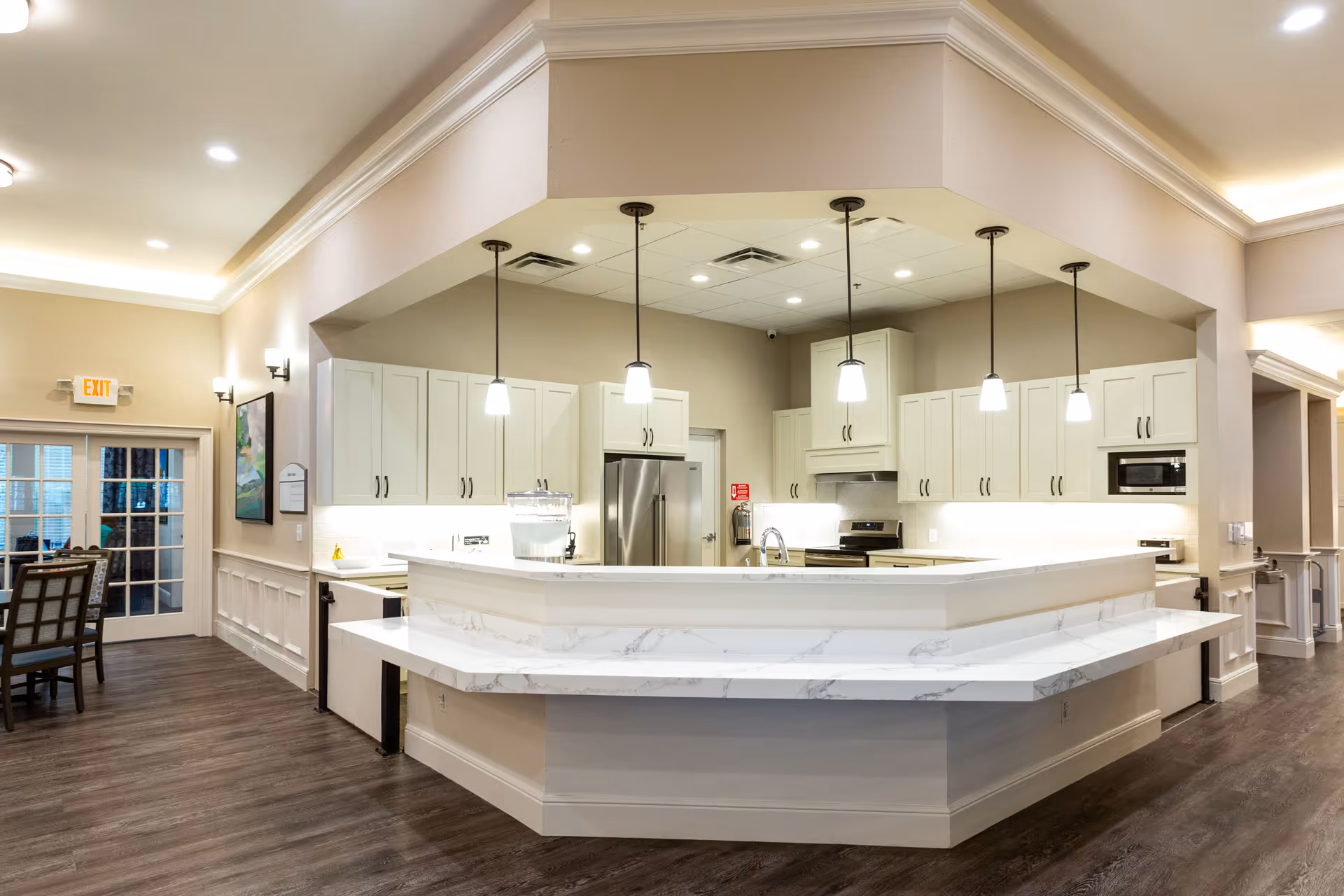Bright and modern kitchen area in an assisted living facility with white cabinetry, stainless steel refrigerator and stove, marble countertops, pendant lights hanging from the ceiling, and wood flooring. Adjacent dining area with chairs and a glass door is visible on the left.