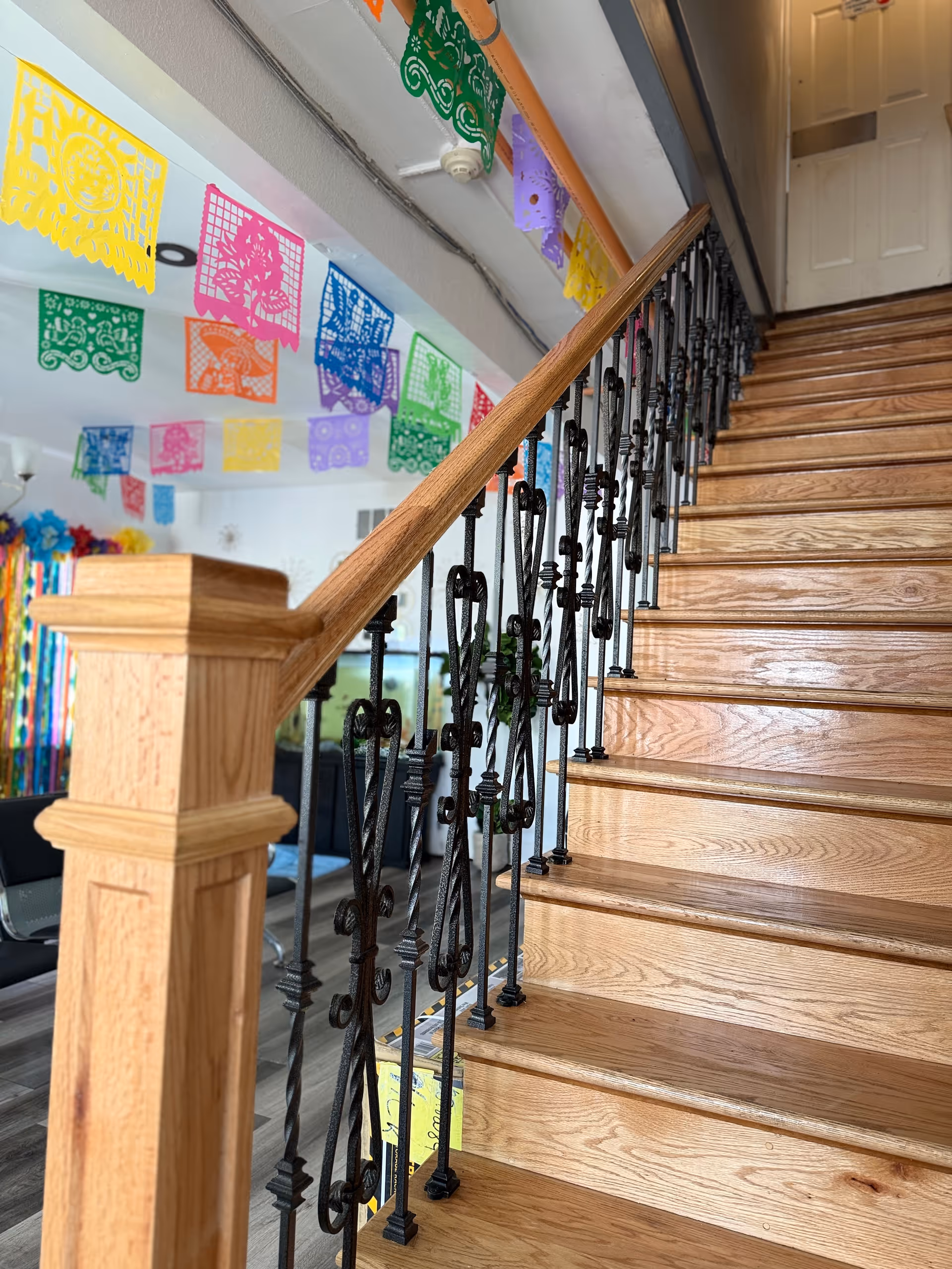 Wooden staircase with wrought-iron balusters rising from a colorful common area decorated with hanging papel picado banners.
