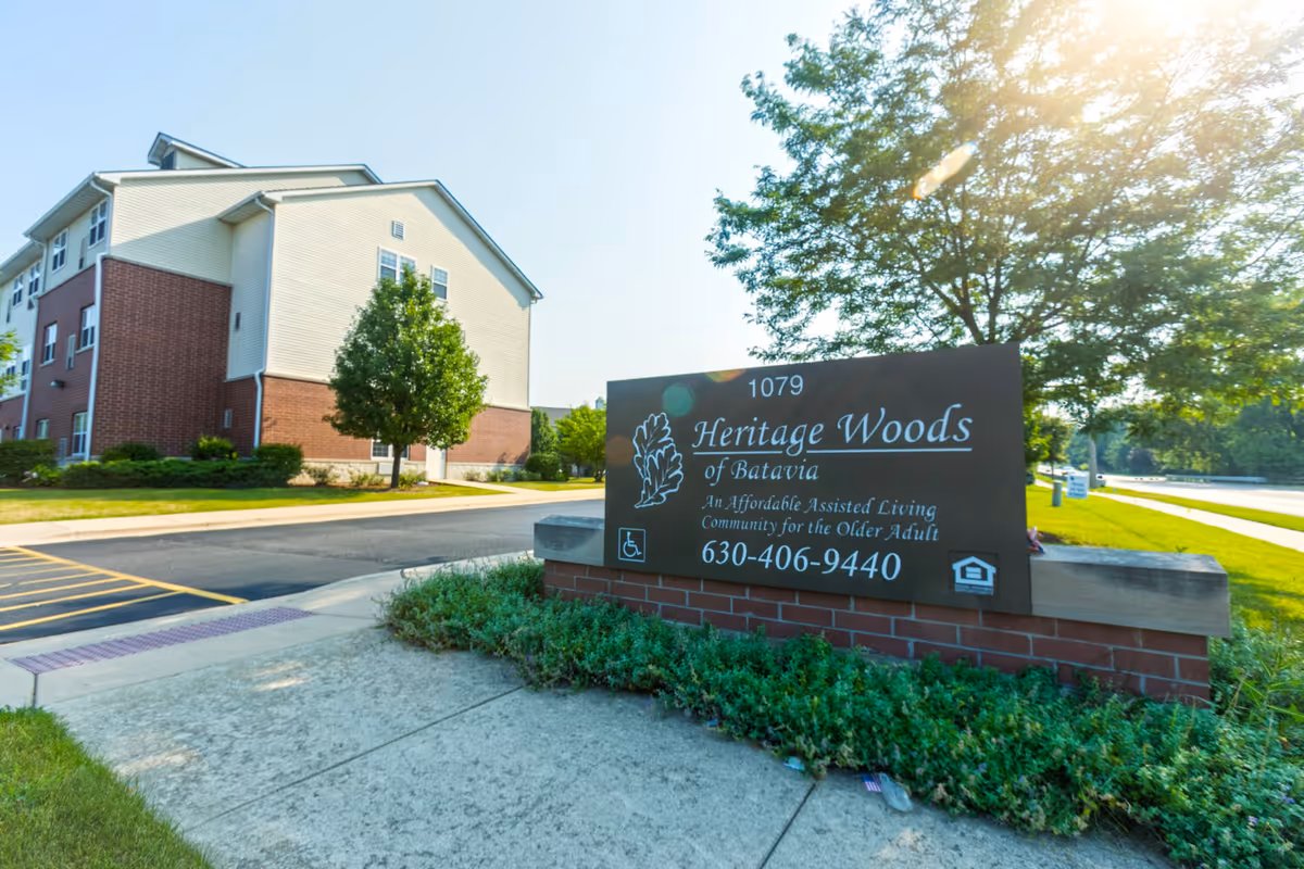 Exterior view of Heritage Woods of Batavia assisted living facility with a large sign in the foreground displaying the facility name, contact number, and accessibility information, surrounded by greenery and a sunny sky.