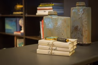 Close-up view of a table with three stacked books titled 'Live Laugh Love' and a decorative pen resting on top. In the background, there are two large ceramic vases and a bookshelf with various books and decorative items.