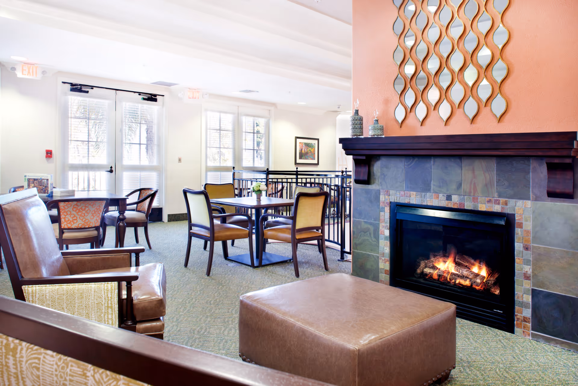 A cozy common area in a senior living facility featuring a lit fireplace with a decorative tile surround and a wooden mantel. Above the fireplace is a wall decoration with multiple wavy mirrors. The room has several tables and chairs arranged for socializing or dining, with large windows and glass doors allowing natural light to fill the space.