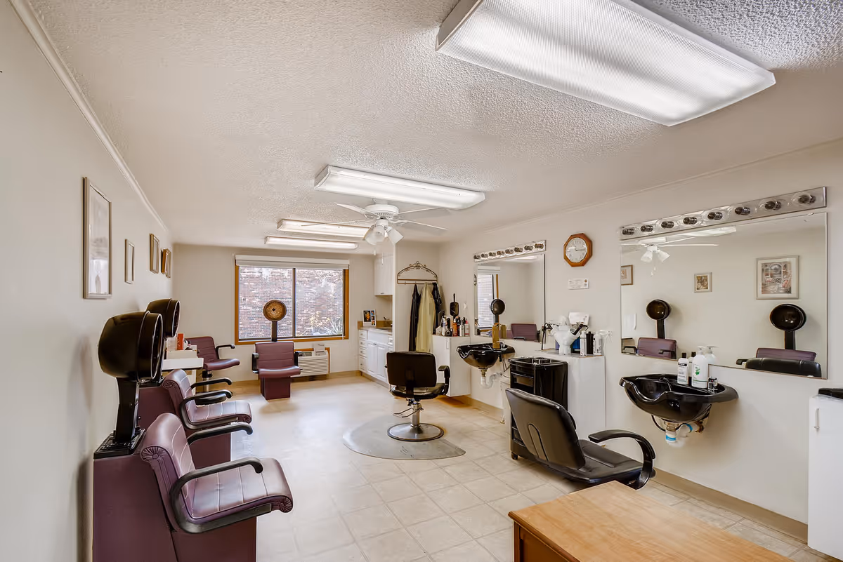 Interior salon with multiple styling chairs, hooded hair dryers, wash basins and large mirrors in a senior living community.