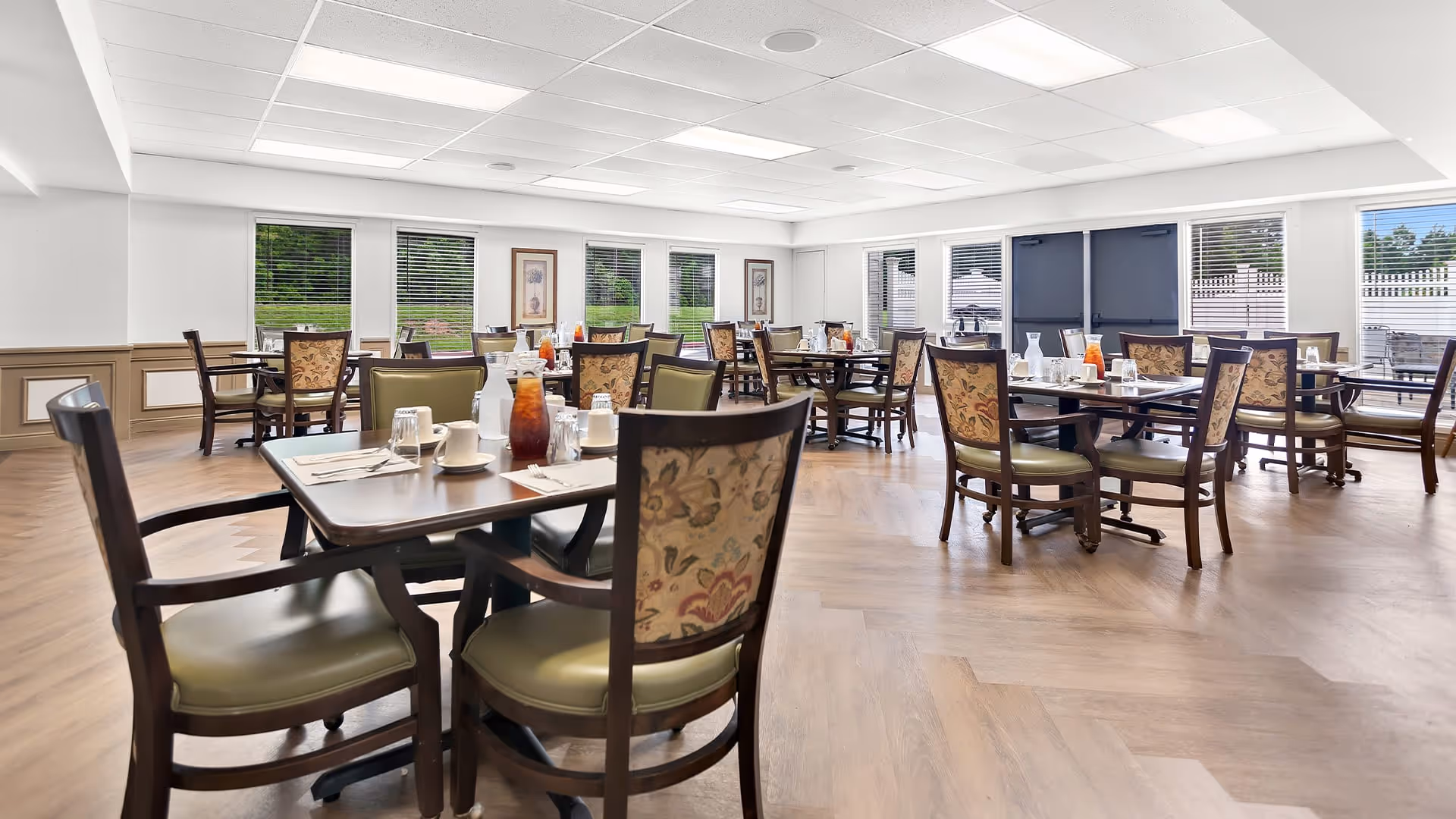 A bright dining room with multiple tables and chairs arranged neatly. Each table is set with cups, glasses, utensils, and pitchers of iced tea. Large windows with blinds allow natural light to fill the room, and framed artwork is displayed on the walls. The floor has a wood pattern, and there are double doors leading outside.