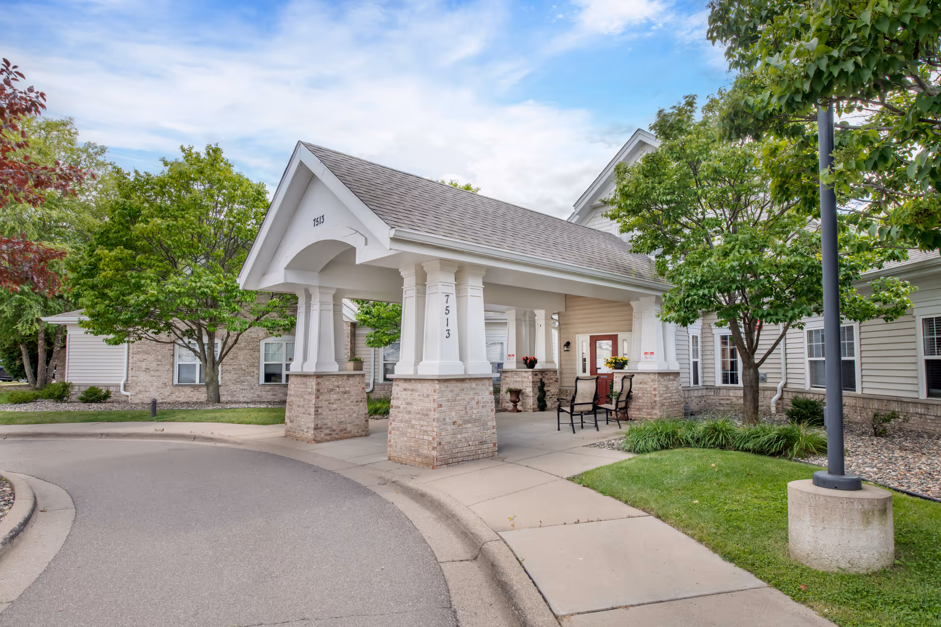 Covered porte-cochere entrance of the Brookdale Eden Prairie building with columns, outdoor seating, and surrounding trees.