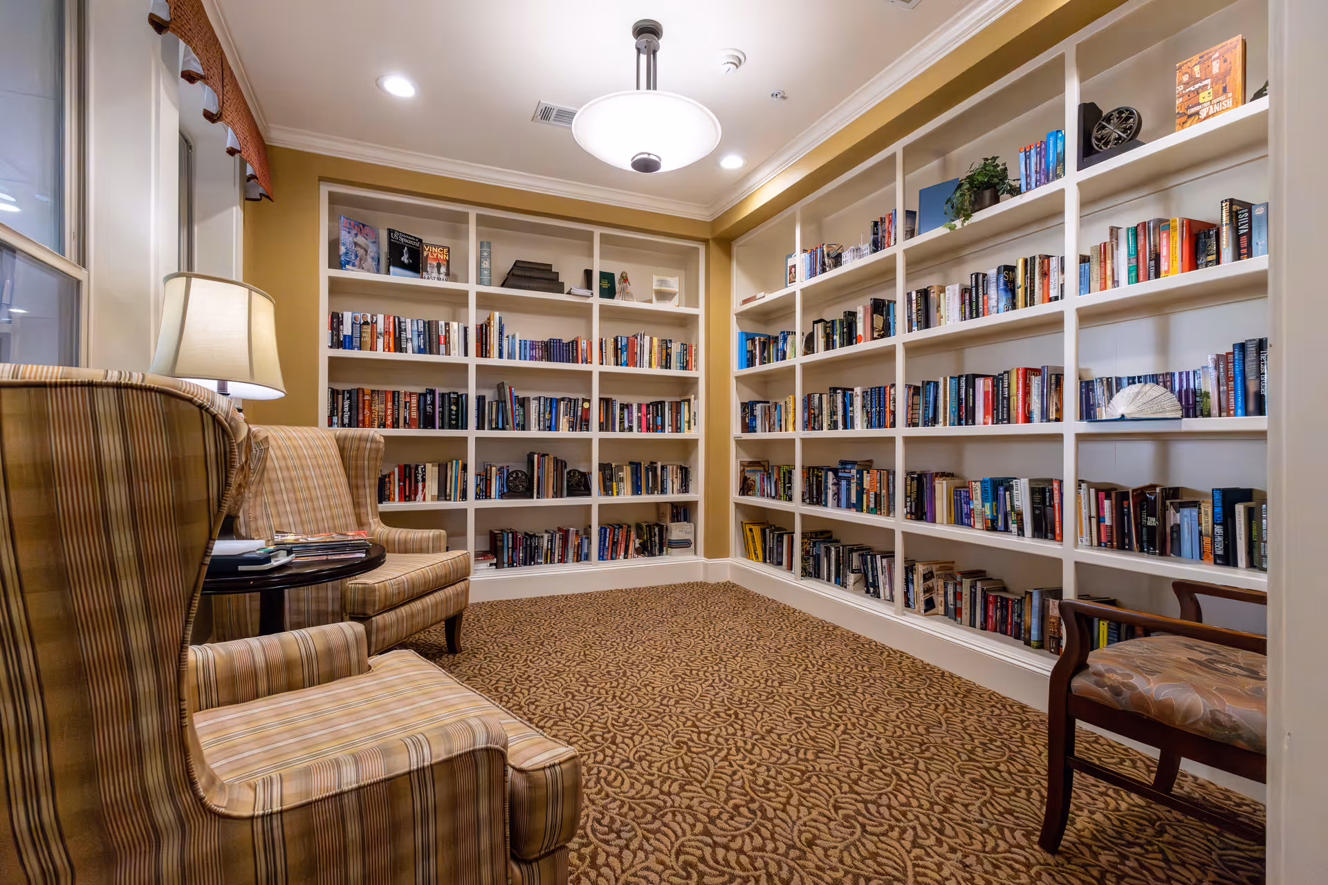 Cozy reading room with built-in white bookshelves filled with books, two striped upholstered armchairs, a wooden chair with patterned cushion, a round side table with a lamp, and a ceiling light fixture. The room has patterned carpet and warm yellow walls.