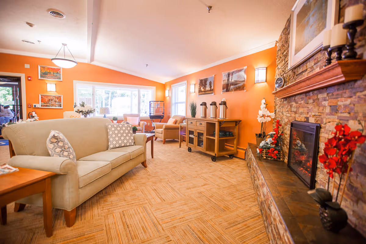 A cozy living room area with orange walls, a beige sofa with patterned cushions, a stone fireplace decorated with red and white flowers, and a wooden sideboard with coffee dispensers. Large windows allow natural light to fill the room, and framed artwork hangs on the walls.