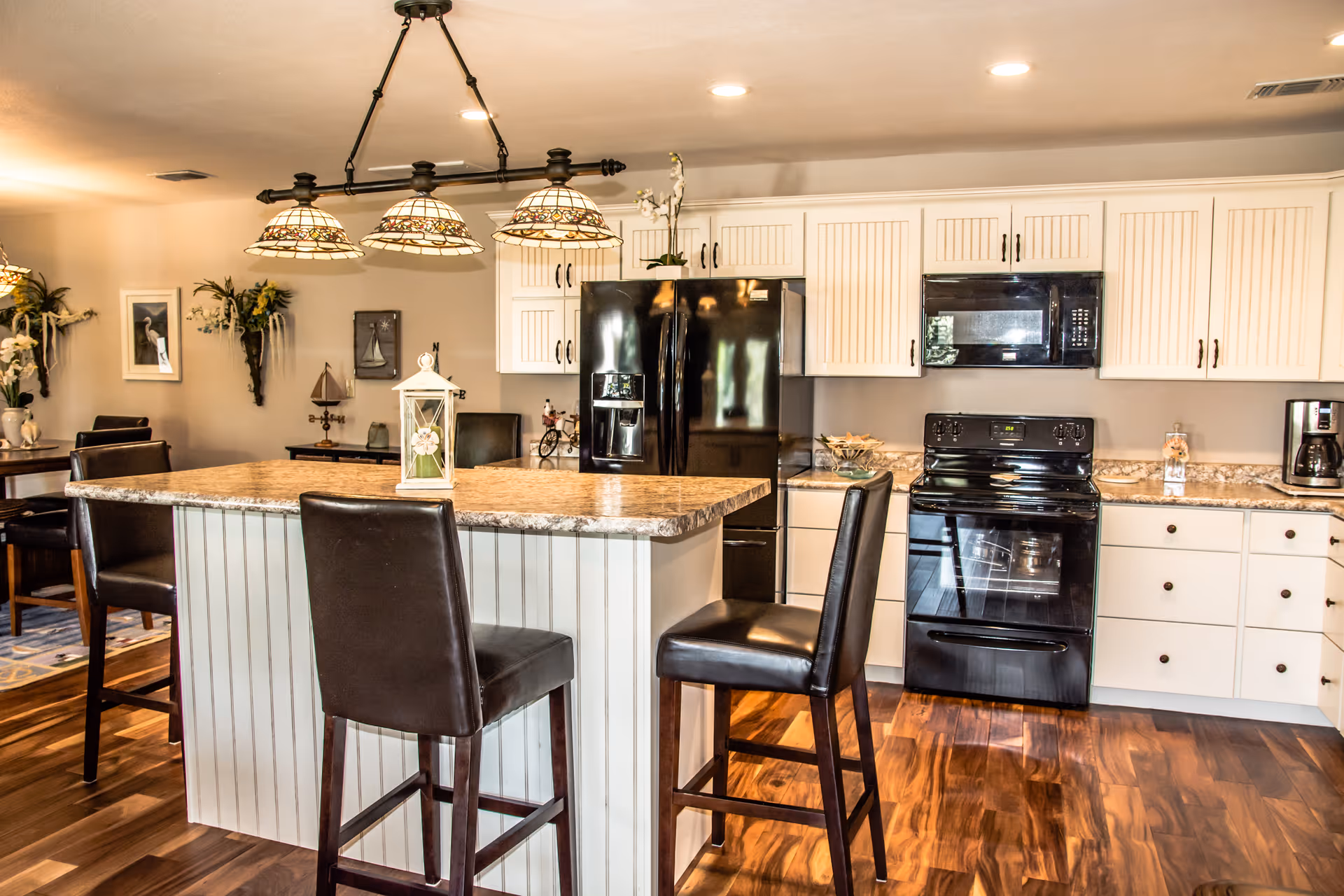 A modern kitchen with white cabinetry, black appliances including a refrigerator, stove, and microwave. There is a kitchen island with a granite countertop and three dark leather chairs. The floor is wooden, and there are decorative wall hangings and a dining area visible in the background.