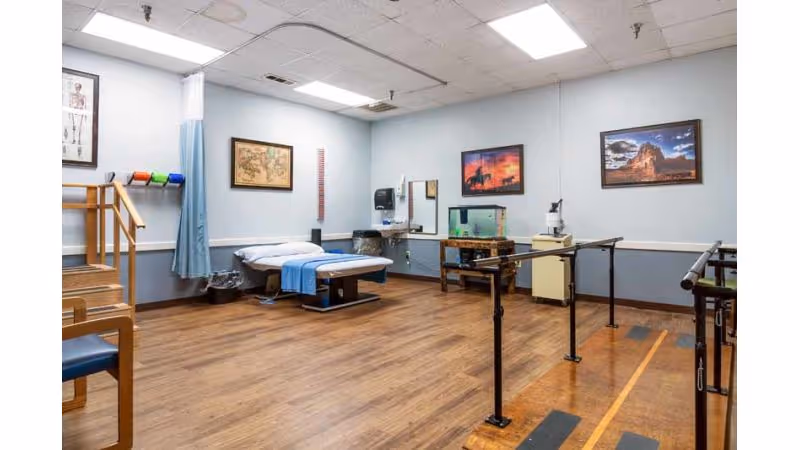 A physical therapy room with wooden flooring, parallel bars for walking exercises, a treatment table with a blue sheet, a small staircase with handrails, and framed pictures on the light blue walls. There is also a fish tank on a wooden stand and medical equipment on a counter.