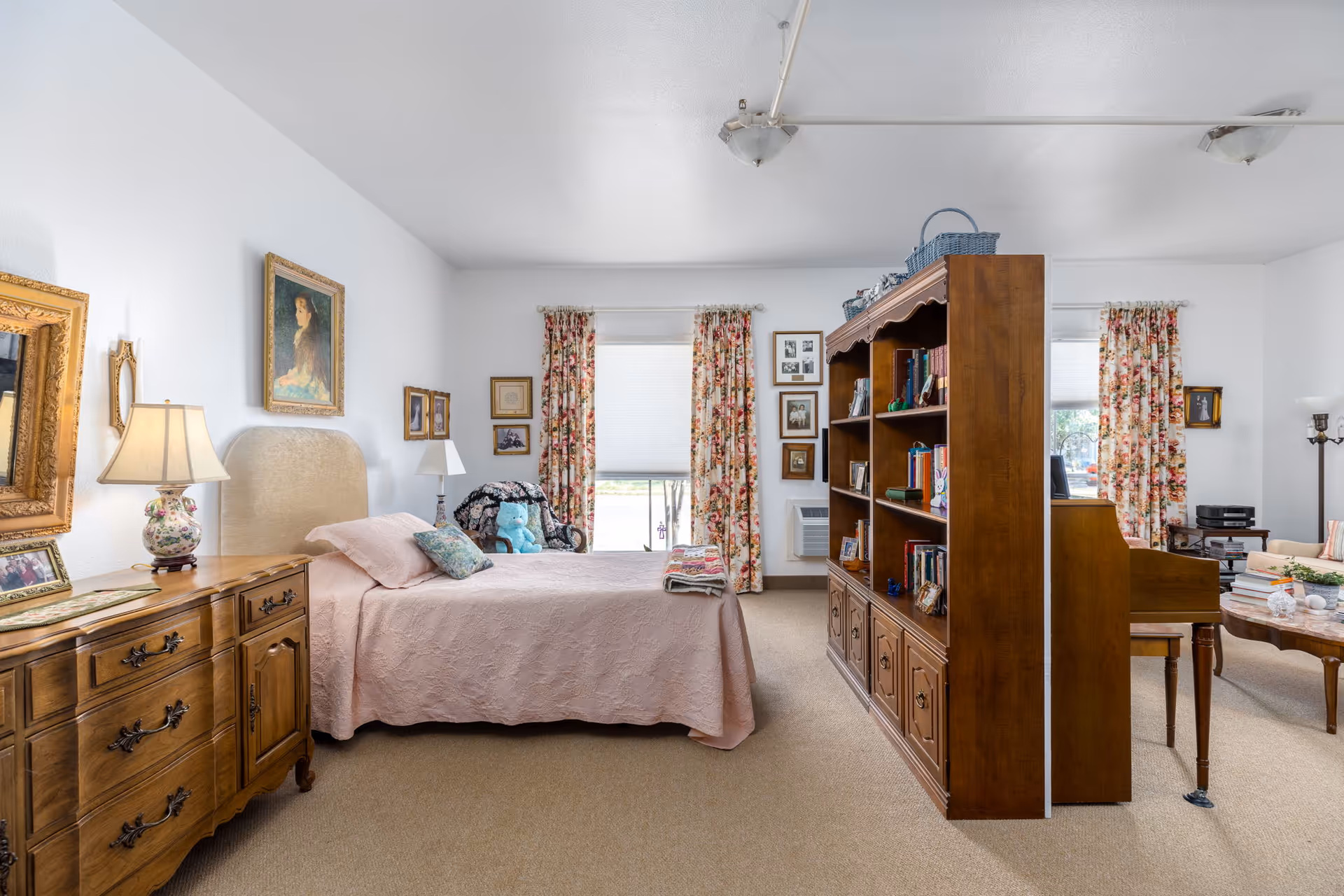 A cozy room in a retirement residence featuring a bed with a pink quilt, a wooden dresser with a floral lamp, and a wooden bookshelf divider separating the sleeping area from a sitting area with a piano and coffee table. The room has floral curtains on two windows and various framed pictures on the walls.