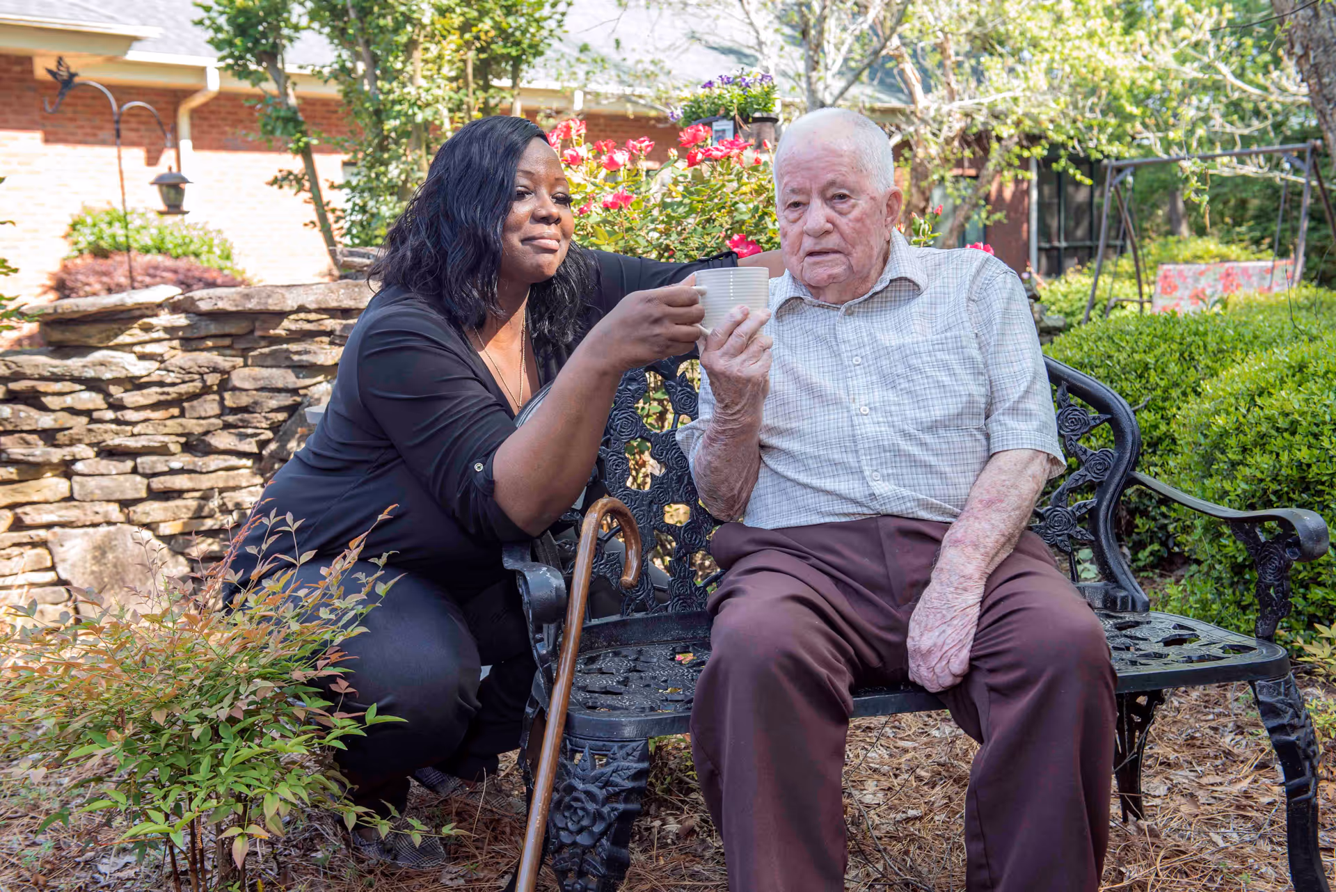 An elderly man sitting on a black metal bench in a garden area, holding a white cup. A woman kneels beside him, smiling and holding another white cup as if toasting. There are green bushes, flowers, and a stone wall in the background.