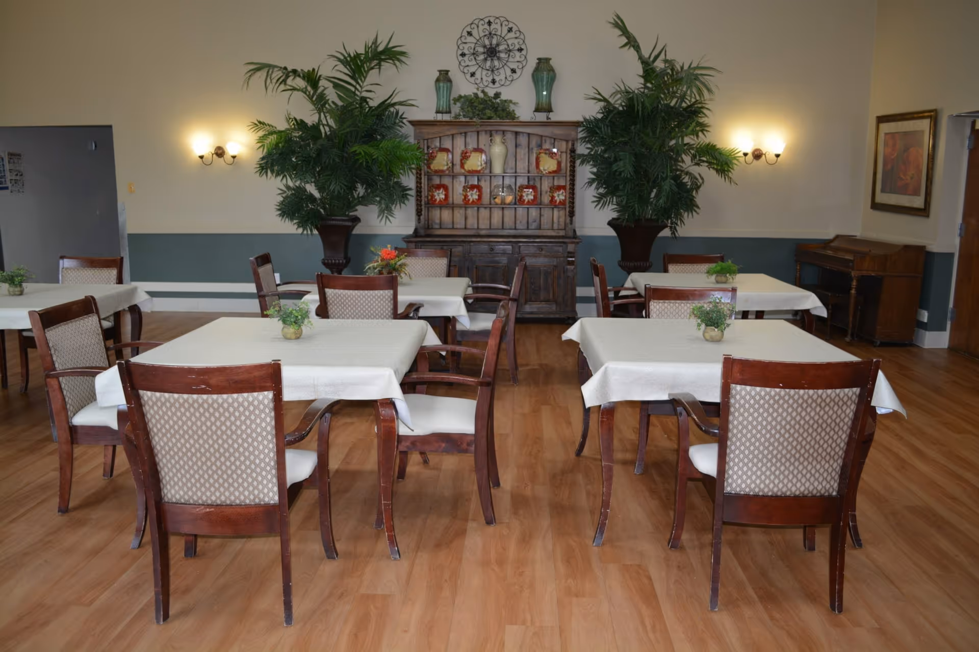Dining area with several tables covered in tablecloths, wooden chairs, potted plants, and a wooden hutch against the back wall.