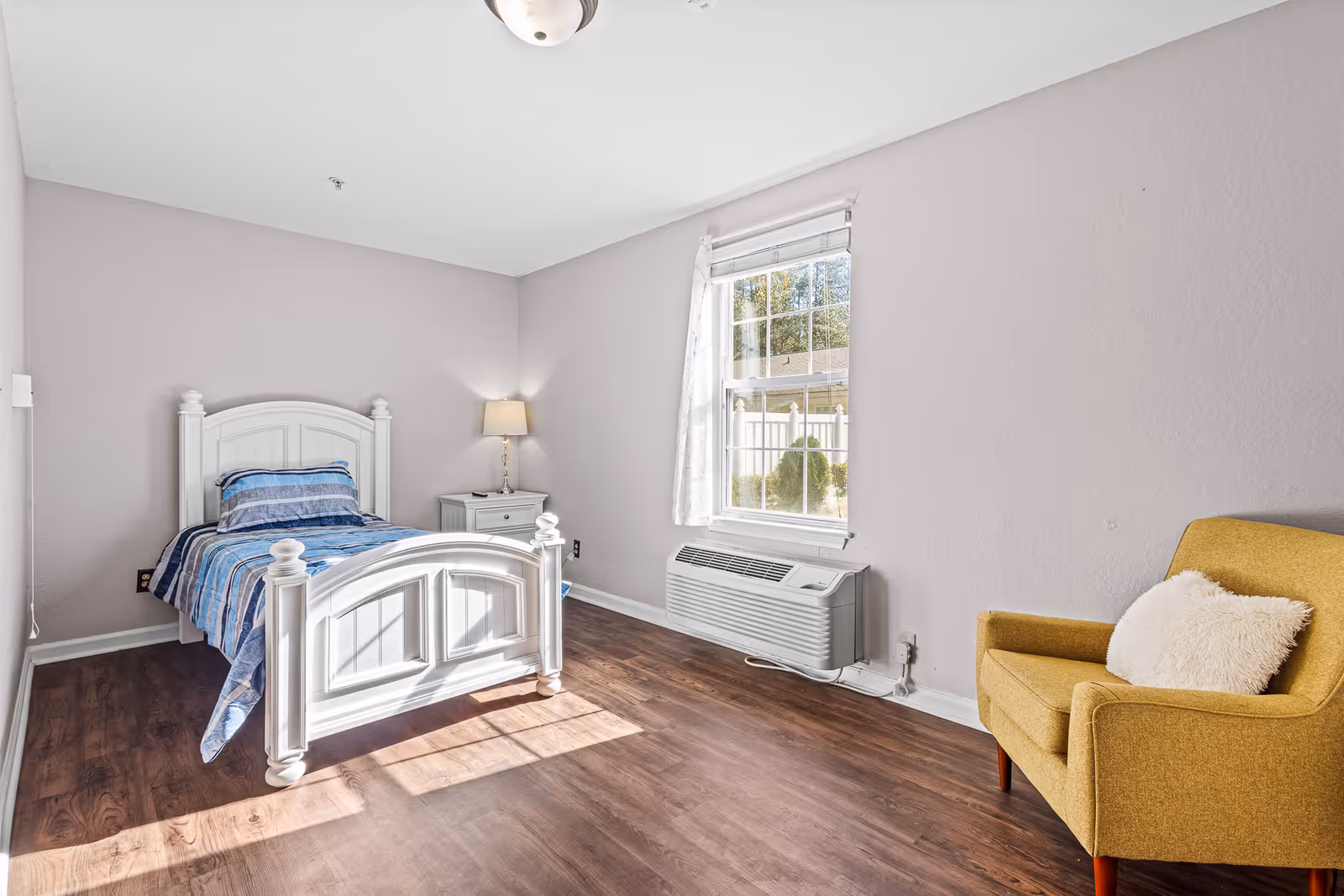 Sunlit bedroom with a white twin bed, nightstand and lamp, a window with an AC unit, and a yellow armchair.