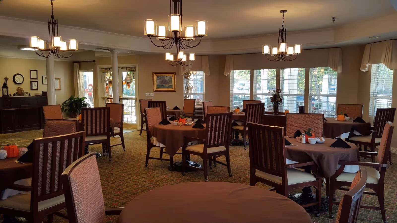 A dining room with several round tables covered with brown tablecloths and set with black napkins, cups, and small decorative pumpkins. The room has multiple wooden chairs with cushioned seats and backs. Large windows with white blinds allow natural light to enter. Three chandeliers hang from the ceiling, illuminating the room. In the background, there is a reception desk with a person seated behind it, a clock on the wall, and framed pictures.