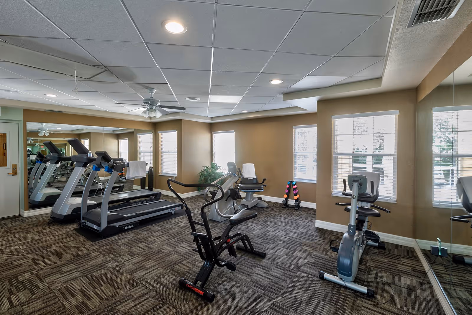 A small fitness room with exercise equipment including two treadmills, a recumbent bike, an upright stationary bike, and a set of colorful dumbbells. The room has large windows with blinds, beige walls, a ceiling fan, and a large mirror covering one wall.