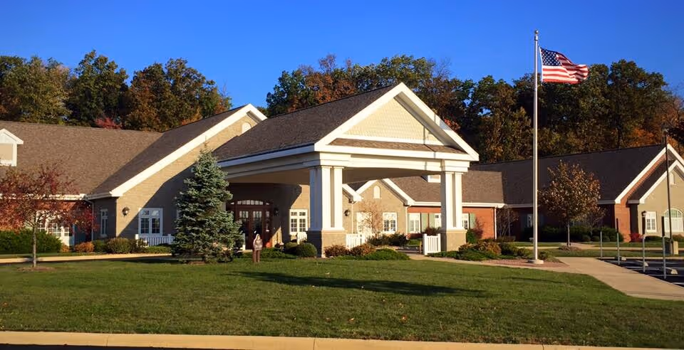 Exterior view of The Inn at Ashland Woods building with a covered entrance, well-maintained lawn, trees, and an American flag on a flagpole against a clear blue sky.