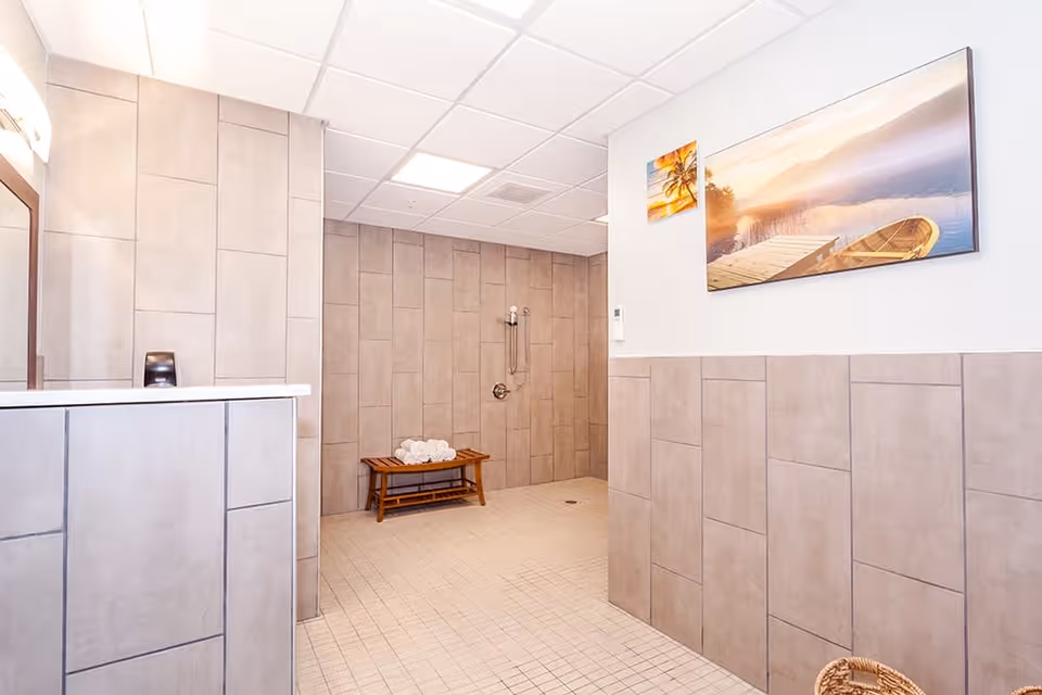 A spacious tiled shower area with beige tiles on the walls and floor. There is a wooden bench with rolled white towels placed on it. The shower has a handheld showerhead attached to the wall. On the right wall, there are two framed pictures depicting serene water and sunset scenes. The ceiling has recessed lighting and a vent.