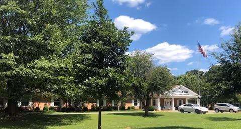 Front view of a brick senior living facility with a portico entrance, American flag, parked cars, and trees on a sunny day.