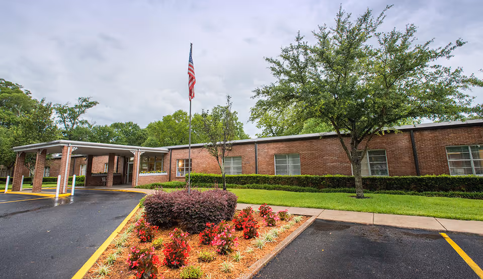 Exterior view of Ashland Place Health and Rehabilitation, LLC showing a single-story brick building with a covered entrance, an American flag on a flagpole, landscaped flower beds with red flowers, green grass, and trees under a cloudy sky.