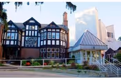 Exterior view of a senior living facility featuring a Tudor-style building with multiple windows and a brick facade, alongside a modern white building with a sloped metal roof entrance and stairs leading up to it.