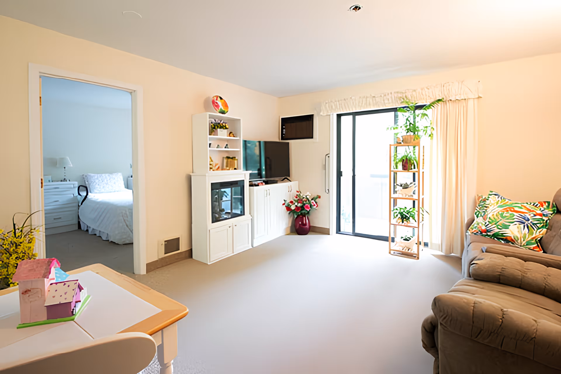 A bright living room with a beige recliner sofa adorned with colorful tropical-patterned pillows. There is a white entertainment unit with a TV and decorative items, a tall plant stand with multiple green plants near a sliding glass door with cream curtains. A doorway leads to a bedroom with a white bed and nightstand. A small table with a pink and purple toy house is in the foreground.