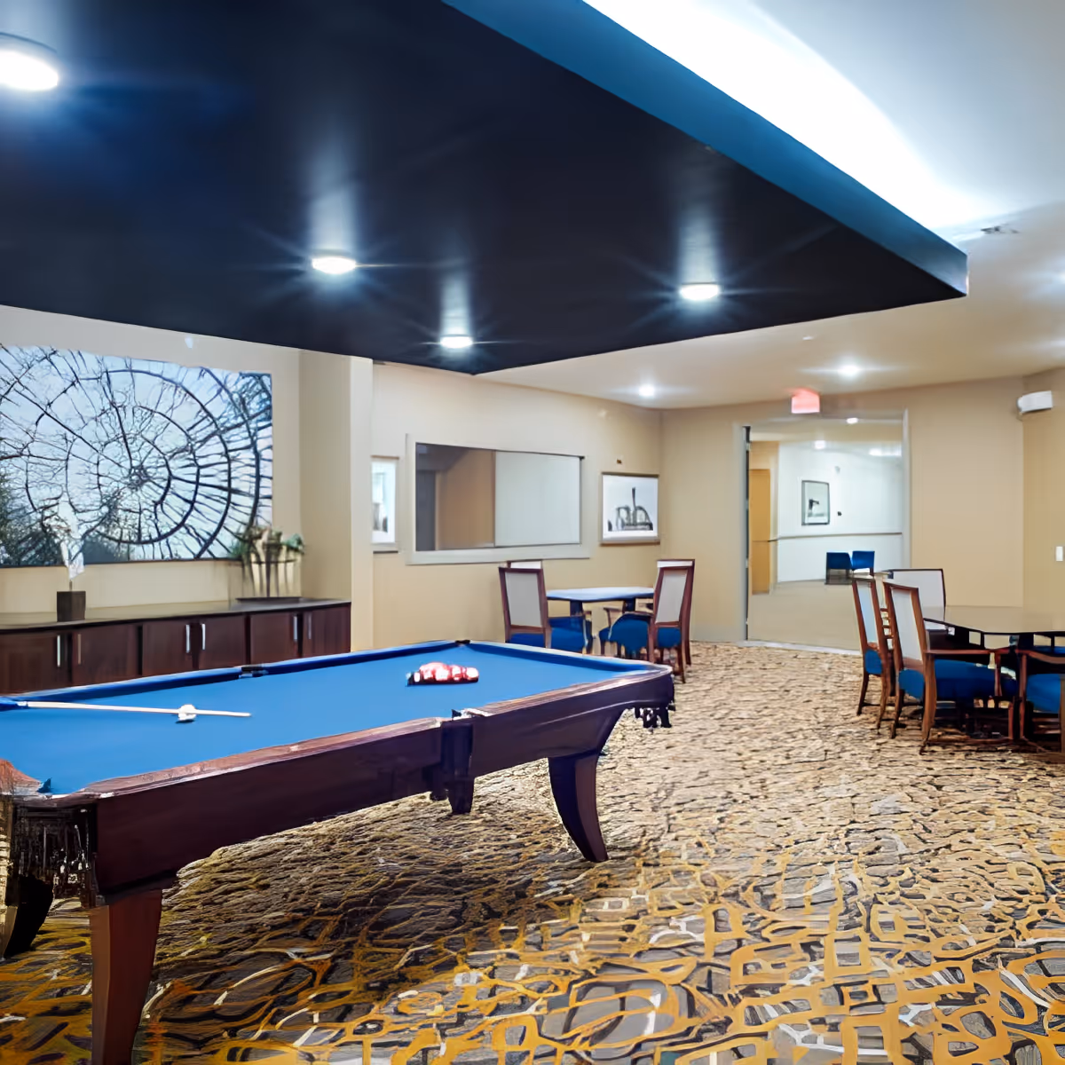 Interior view of a recreational room featuring a pool table with a blue felt surface and a set of billiard balls arranged on it. The room has patterned carpet flooring, beige walls, and a dark ceiling panel with recessed lighting. There are several tables and chairs with blue cushions along the walls, and framed artwork is displayed on the walls.