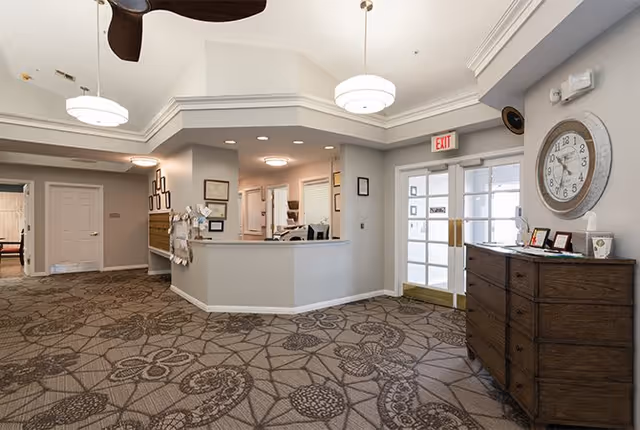 Interior view of a senior living facility reception area with a front desk, patterned carpet, a large wall clock, and a wooden dresser. The space is well-lit with ceiling lights and has an exit door with glass panels.