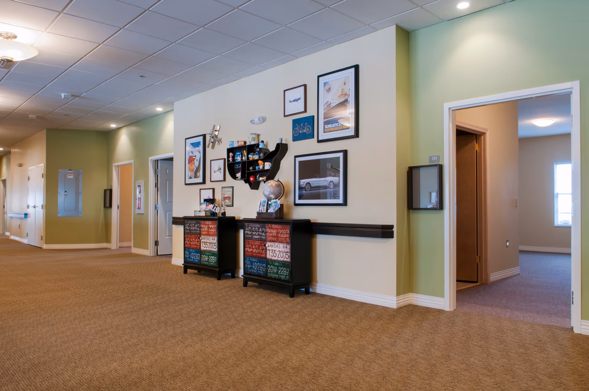 Wide carpeted hallway in a senior living facility featuring decorative cabinets, framed pictures on a light green wall, and open doorways to rooms.
