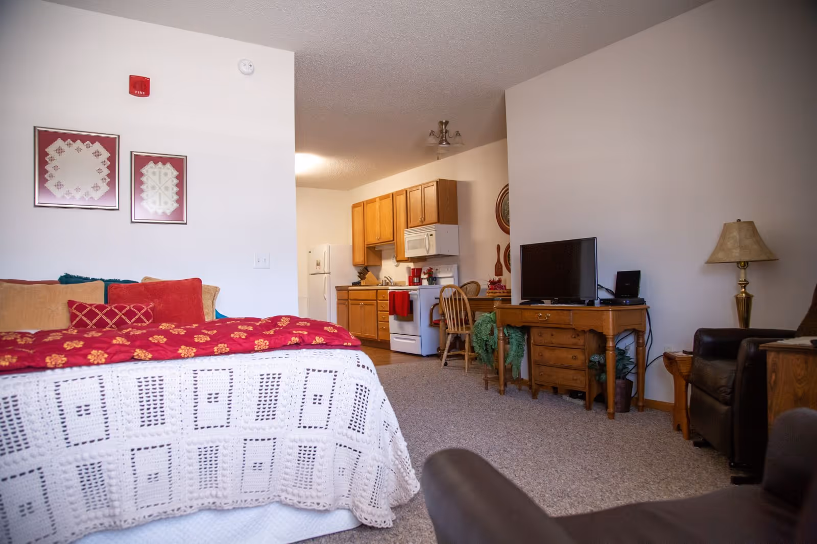 Interior view of a senior living facility room featuring a bed with a white crocheted blanket and red floral quilt, a small kitchen area with wooden cabinets, a white refrigerator, stove, and microwave, a wooden desk with a TV on top, a wooden chair, and a lamp beside a dark armchair. The walls are decorated with framed artwork and the floor is carpeted.