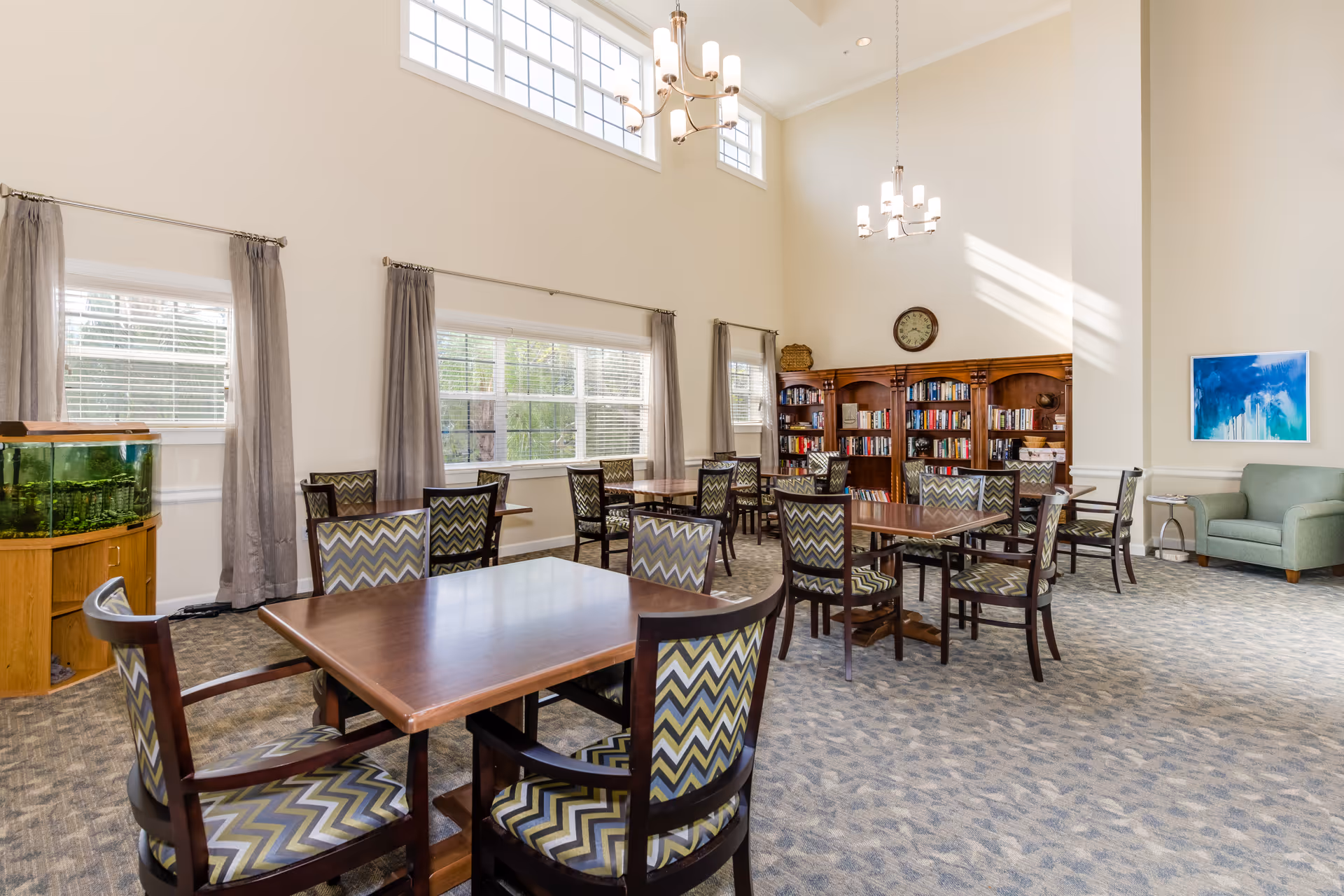 A bright and spacious common area with multiple wooden tables and chairs featuring zigzag patterned upholstery. The room has large windows with gray curtains, high ceilings with two chandeliers, a wooden bookshelf filled with books, a wall clock, a green armchair, a side table, and a fish tank in the corner.