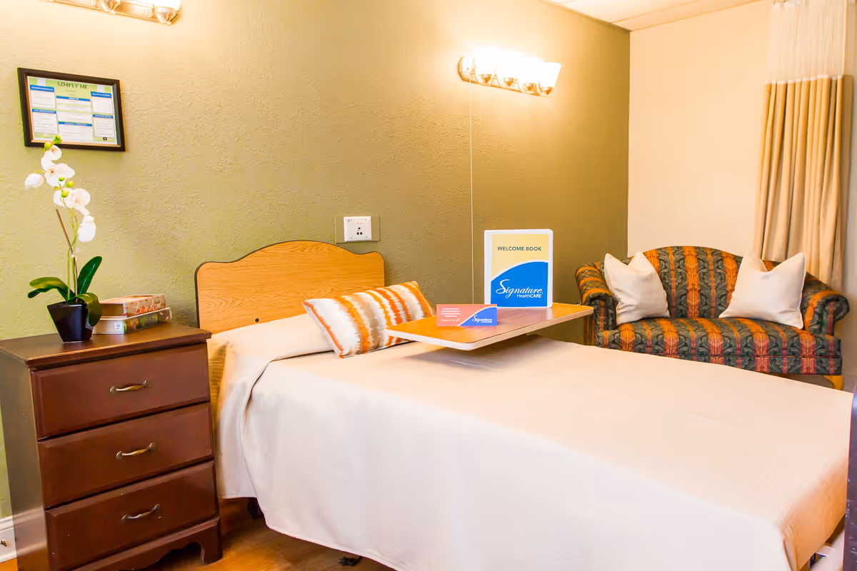 A neatly made single bed with a wooden headboard in a room. There is a bedside table with three drawers on the left, holding a potted orchid and some books. On the bed is a tray with a welcome book and some cards from Signature HealthCARE. In the background, there is a patterned loveseat with two white pillows and beige curtains on the right side of the room. The walls are painted in two tones, with a green accent wall behind the bed and a light beige wall on the right.