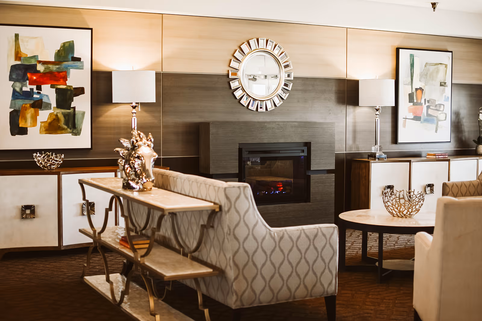 A cozy living room area featuring a modern electric fireplace centered on a dark wood-paneled wall. On either side of the fireplace are white cabinets with decorative items and abstract artwork hanging above. Two table lamps with white shades provide warm lighting. In front of the fireplace are patterned upholstered chairs and a round coffee table with a decorative bowl. A console table with a metallic horse head sculpture is positioned behind one of the chairs.