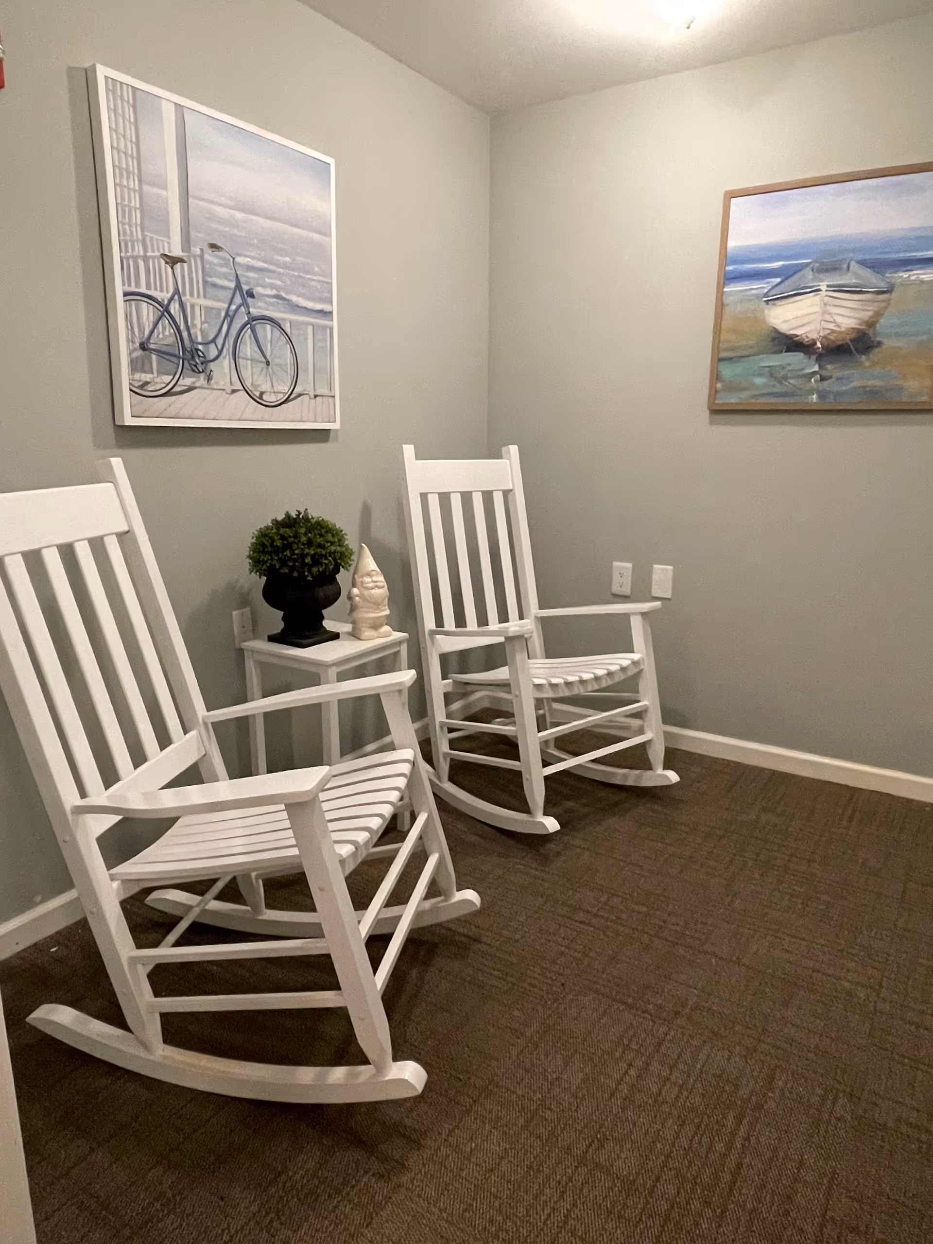 Two white wooden rocking chairs and a small side table with a plant and figurine in a carpeted room with coastal artwork on the walls.