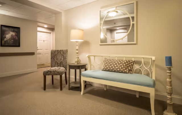 A cozy seating area in a hallway with a white bench featuring a blue cushion and a patterned pillow, a small round side table with a lamp, a patterned upholstered chair, a tall candle holder with a blue candle, and a round mirror on the wall reflecting the ceiling light and door.
