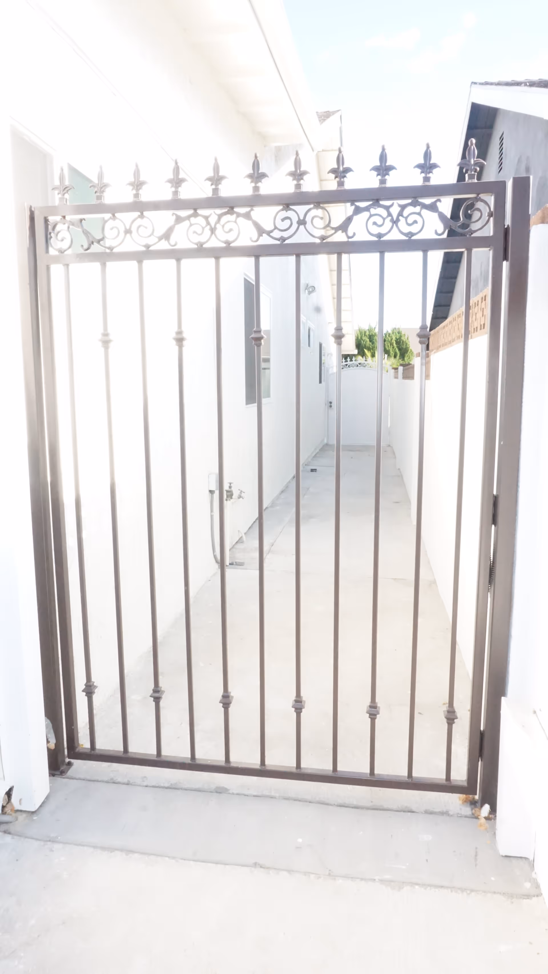 A black metal gate with decorative fleur-de-lis finials at the top, blocking a narrow concrete pathway between two white buildings. The pathway leads to a white wall with a smaller gate in the distance, and there are some green trees visible beyond the wall.