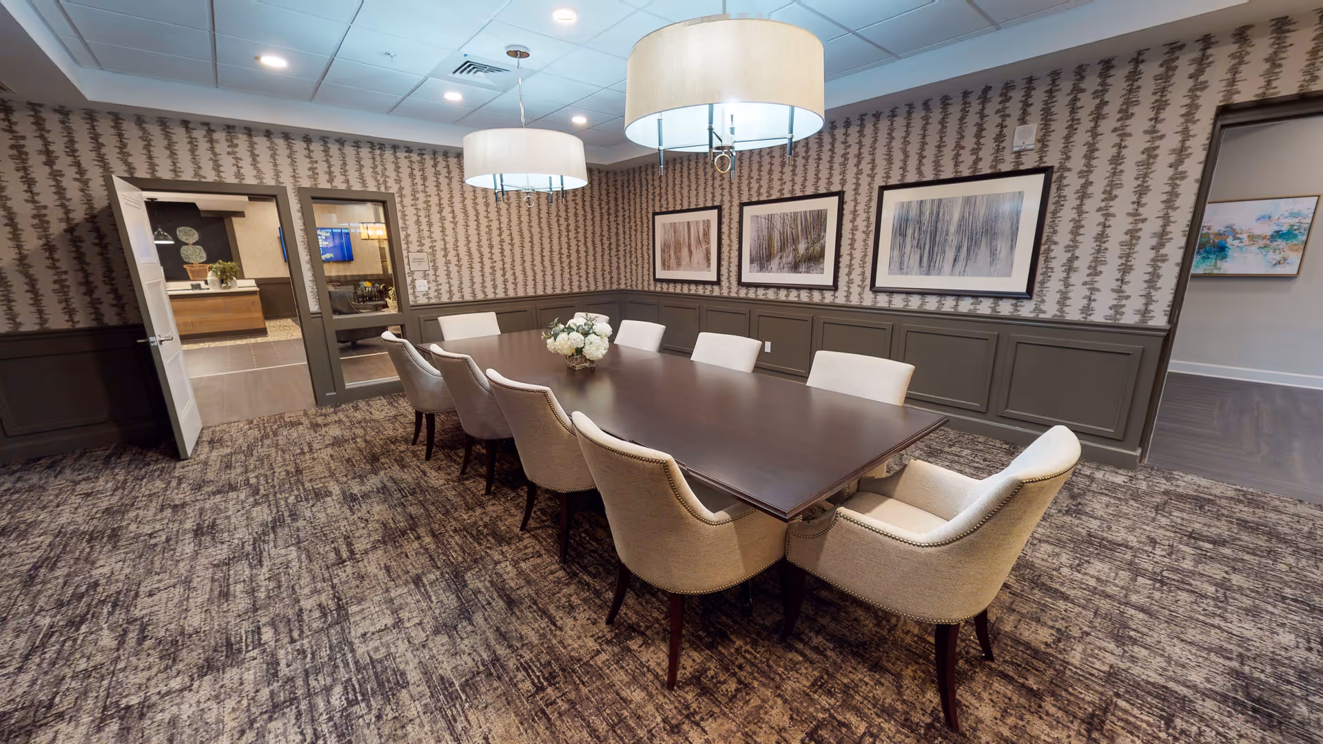 A well-lit conference or dining room with a long dark wooden table surrounded by eight beige upholstered chairs. The room features patterned wallpaper, three framed abstract art pieces on the wall, and two large round pendant lights hanging from the ceiling. The floor is carpeted with a textured design, and there is an open door leading to another room.