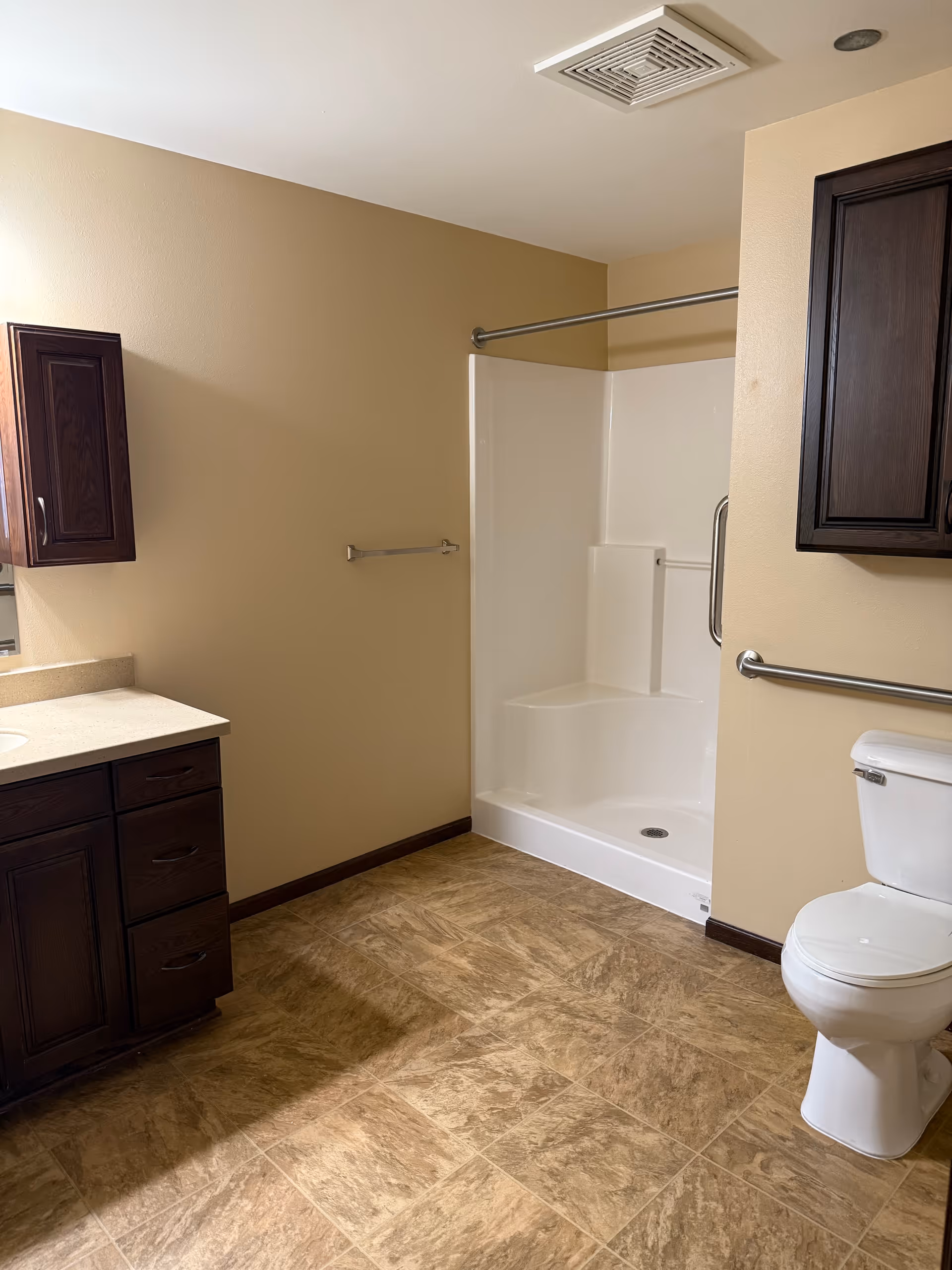 A bathroom with beige walls and brown tiled floor. It features a white toilet with a metal grab bar next to it, a white shower with built-in seating and a metal grab bar, and dark wood cabinets with a beige countertop and sink.