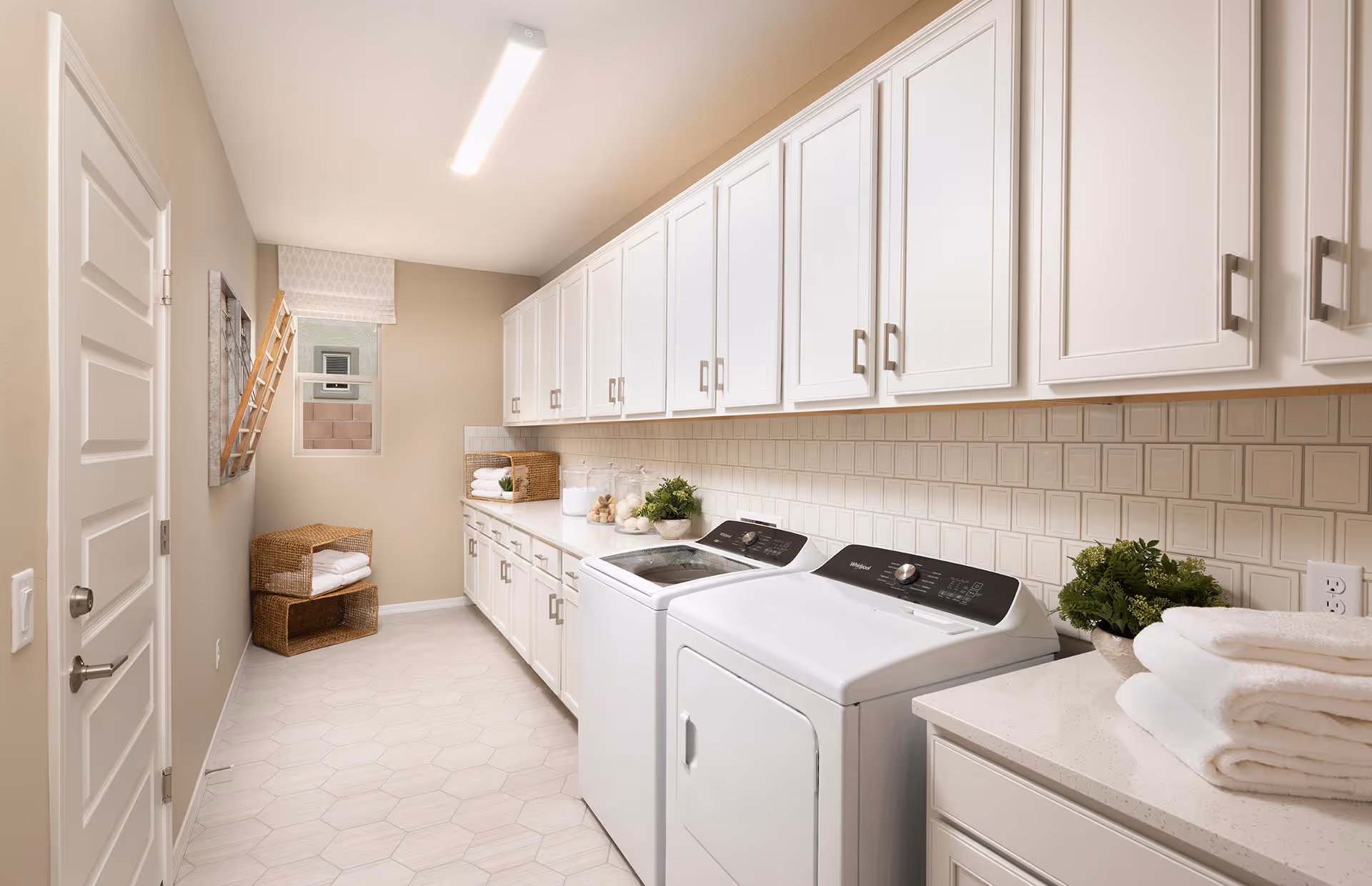 Bright laundry room with side-by-side washer and dryer, white cabinets, tile backsplash, and folded towels on the counter.