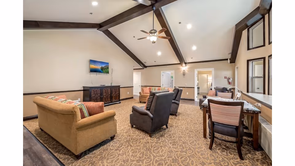 A spacious living room area in The Cottages of Lynnwood featuring beige walls, a high vaulted ceiling with dark wooden beams, a ceiling fan with light, patterned carpet, and multiple seating options including two brown armchairs, a beige sofa with colorful cushions, and a wooden table with chairs near windows.