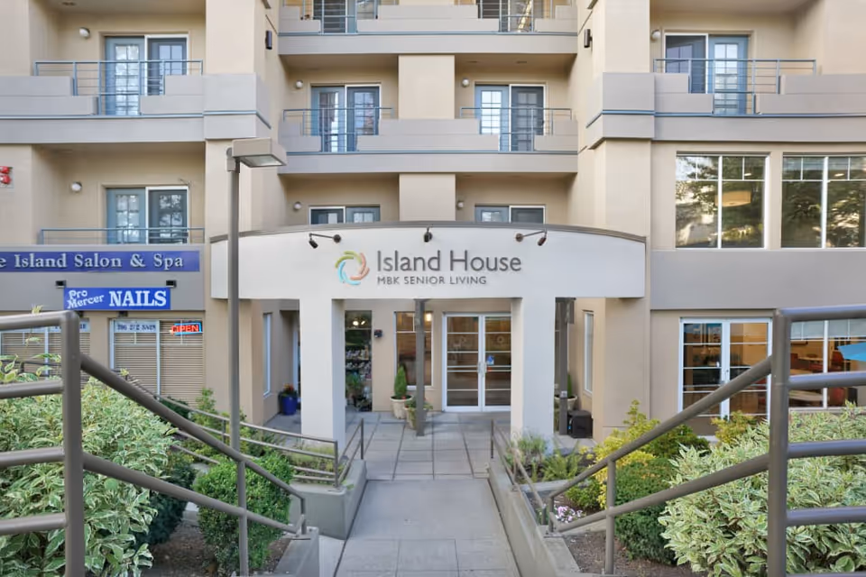 Front entrance of Island House MBK Senior Living facility with a ramp and stairs leading up to glass double doors. The building has multiple balconies and windows. To the left, there is signage for Pro Mercer Nails and The Island Salon & Spa.