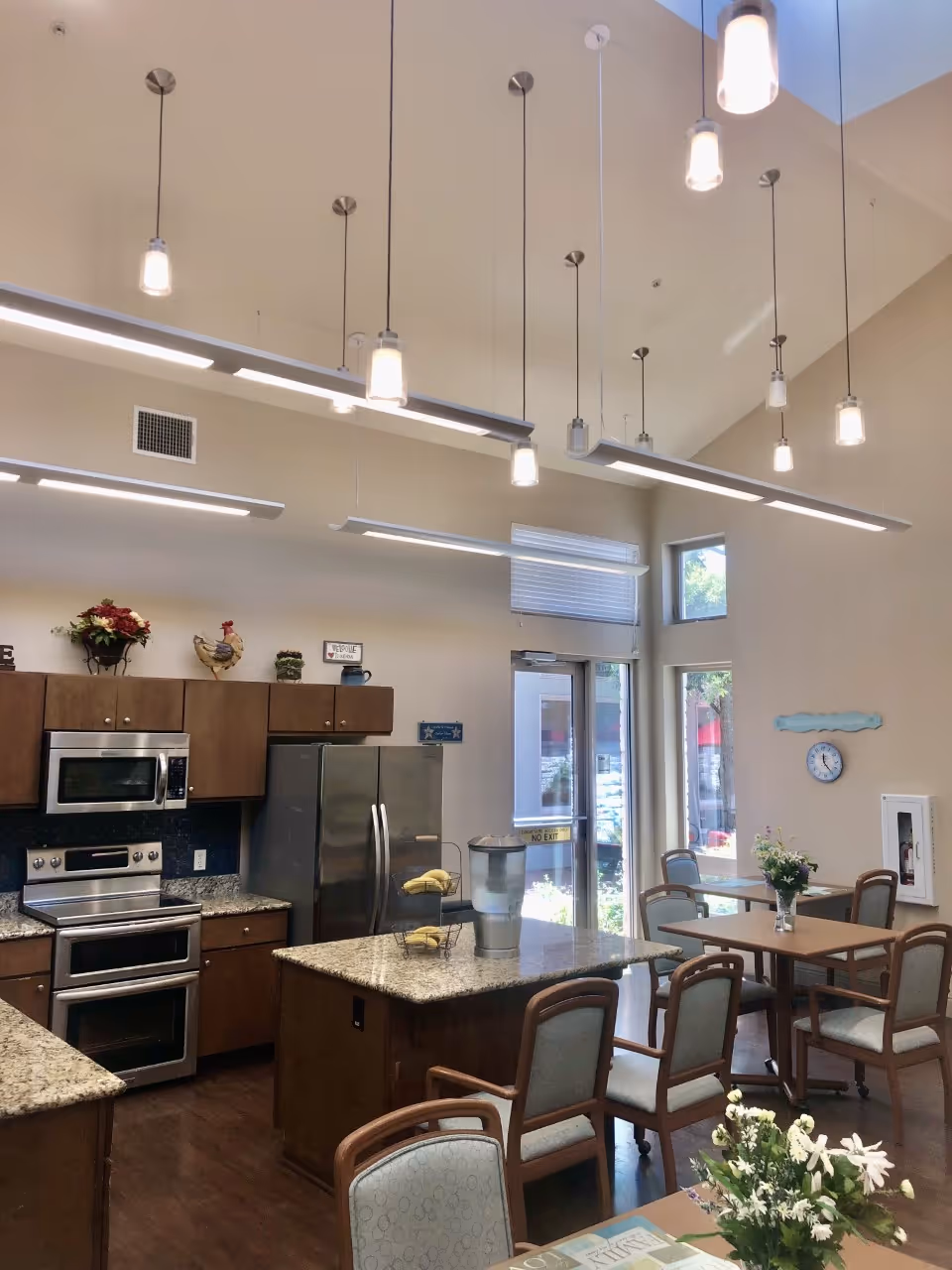A bright kitchen and dining area with high ceilings and multiple hanging lights. The kitchen features stainless steel appliances including a microwave, oven, and refrigerator, with granite countertops and wooden cabinets. There is a kitchen island with a water dispenser and a fruit stand holding bananas. Adjacent to the kitchen are tables and chairs arranged for dining, with flower vases on the tables. Large windows and a glass door allow natural light to enter the space.