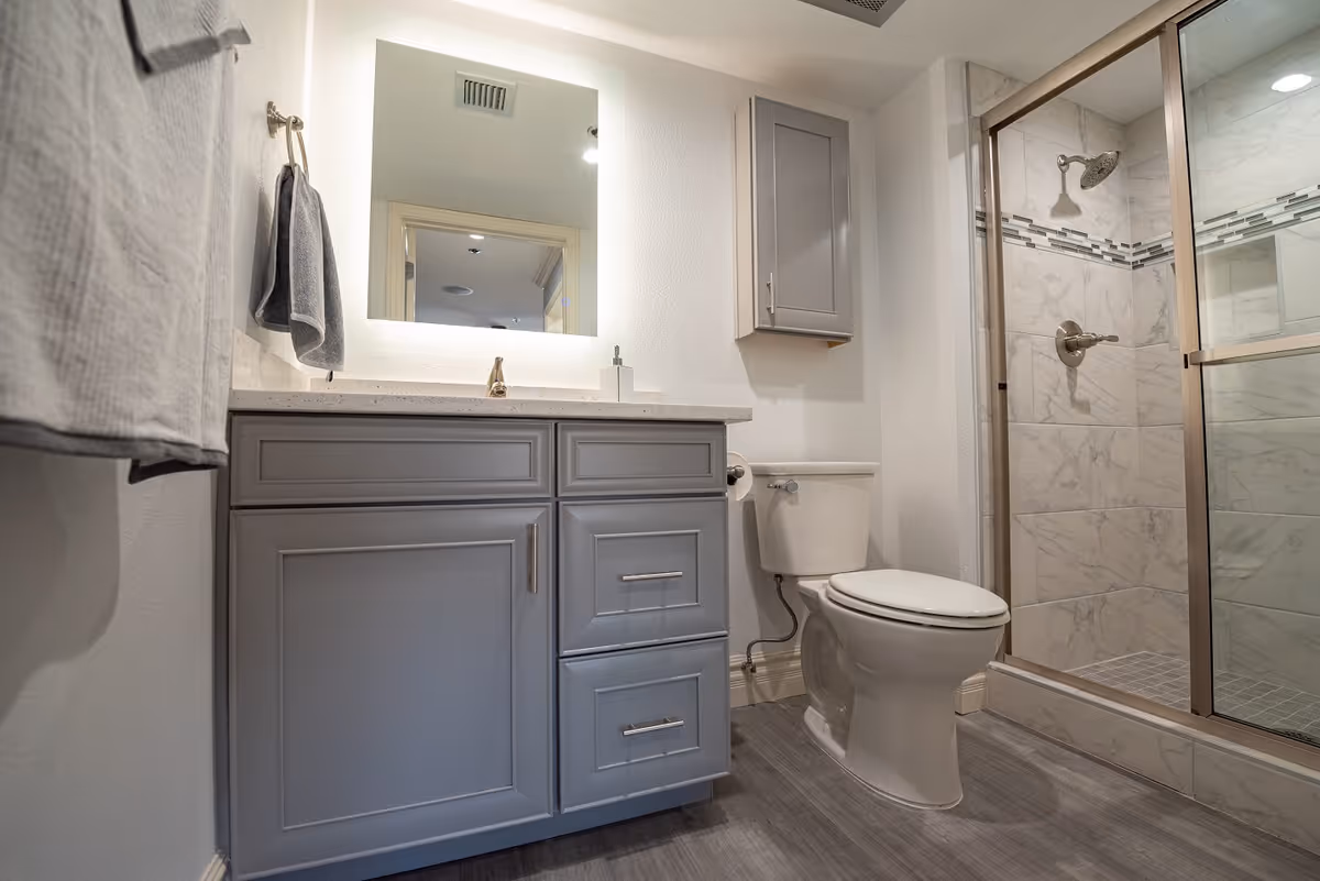 A modern bathroom featuring a gray vanity with a white countertop, a large illuminated mirror above the sink, a toilet, and a glass-enclosed shower with marble-like tiles and a decorative tile border.