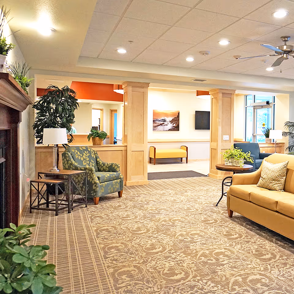 A bright and spacious senior living facility common area with patterned carpet, a yellow sofa with a decorative pillow, a green patterned armchair, side tables with lamps, potted plants, wooden columns, a ceiling fan, and wall art. The area has large windows and a door letting in natural light.