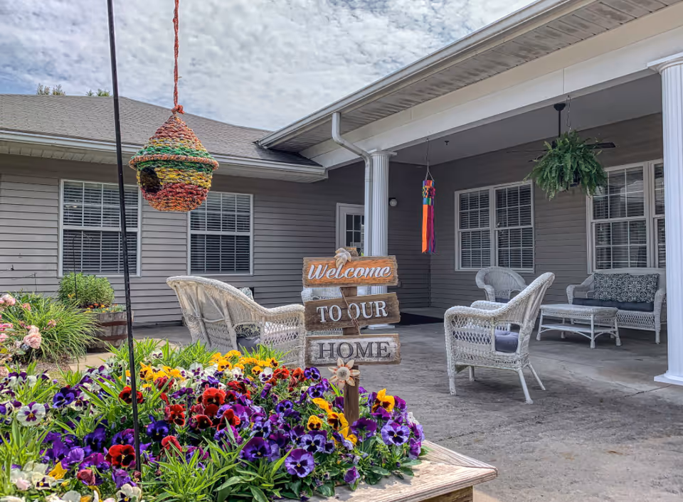 Outdoor patio area with white wicker chairs and a small table under a covered porch. A wooden sign in the foreground reads 'Welcome to our Home' surrounded by colorful flowers. A hanging woven birdhouse and a hanging plant are also visible.