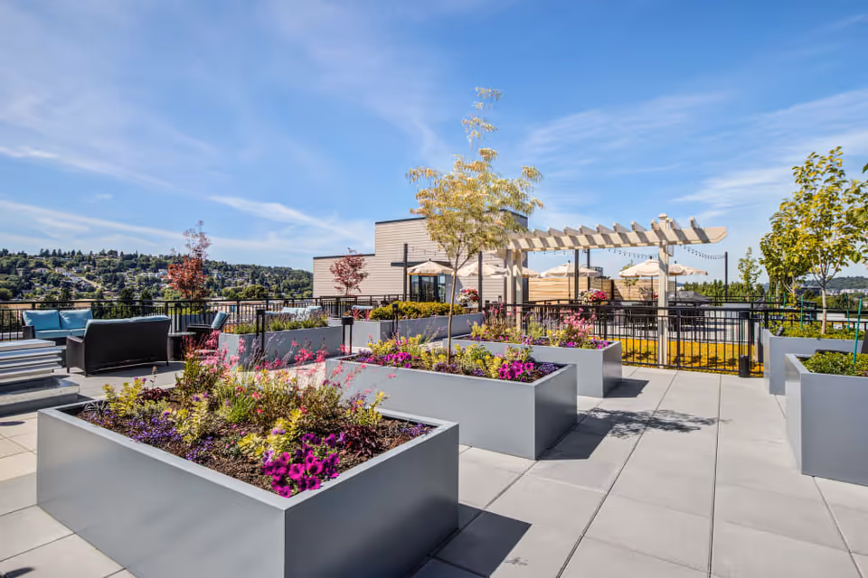 Outdoor patio area with large rectangular planters filled with colorful flowers and plants. There are seating areas with chairs and tables under umbrellas and a pergola. The background shows a scenic view of trees and hills under a blue sky with some clouds.