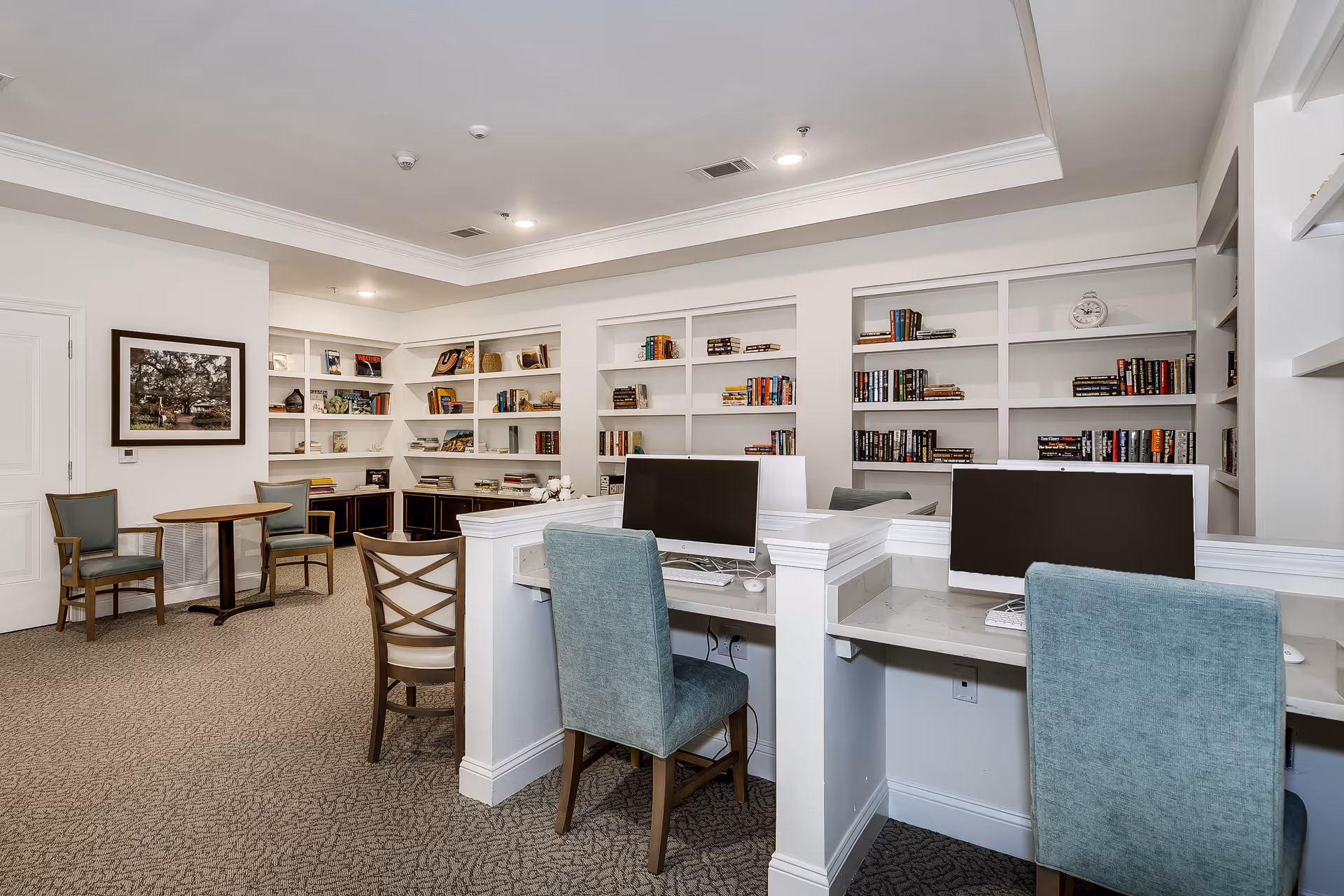 A quiet, well-lit room with built-in white bookshelves filled with books and decorative items. There are two computer workstations with monitors and teal upholstered chairs in the foreground. In the background, there is a small round table with two chairs and a framed picture on the wall. The room has a carpeted floor and a tray ceiling with recessed lighting.