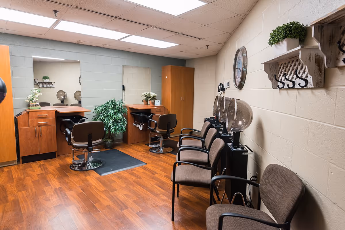 Interior view of a senior living facility's hair salon area featuring wooden flooring, two styling stations with mirrors and chairs, a row of waiting chairs, hair dryers, a wall clock, and decorative plants on shelves and counters.